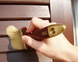 a hand holding a paintbrush varnishing wooden shutters